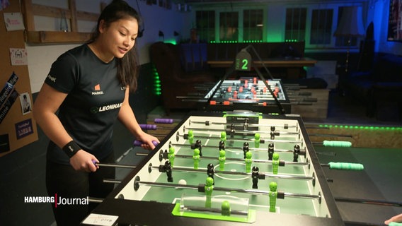 Linh Tran spielt konzentriert Tischfußball in einem Raum mit mehreren Kickertischen und grüner LED-Beleuchtung. © Screenshot 