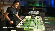 Linh Tran spielt konzentriert Tischfußball in einem Raum mit mehreren Kickertischen und grüner LED-Beleuchtung. © Screenshot 