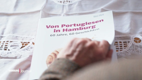 Ein Buch mit dem Titel „Von Portugiesen in Hamburg – 60 Jahre, 60 Geschichten“ liegt auf einer bestickten Tischdecke. © Screenshot 