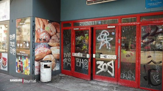 Ein heruntergekommener Laden mit roten, mit Graffiti besprühten Türen. Links Werbeplakate für Backwaren und Getränke. Ein Mülleimer davor. © Screenshot 