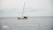 Ein Segelboot mit deutscher Flagge und mehreren Personen an Bord treibt auf ruhigem Wasser unter einem bewölkten Himmel. © Screenshot 