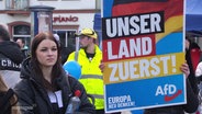 Eine junge Frau steht neben einem Wahlplakat der AfD. © Screenshot 