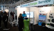 Ein Messestand von „NB Propangas & Energie Service“. Ein Mitarbeiter im blauen Shirt spricht mit einer Frau. Andere Besucher laufen durch die Halle. © Screenshot 