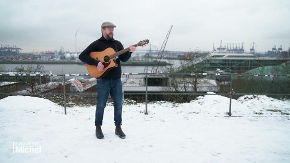 Ein Mann mit Gitarre steht am verschneiten Altonaer Balkon vor der Kulisse des Hamburger Hafens. © Screenshot 