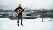 Ein Mann mit Gitarre steht am verschneiten Altonaer Balkon vor der Kulisse des Hamburger Hafens. © Screenshot 