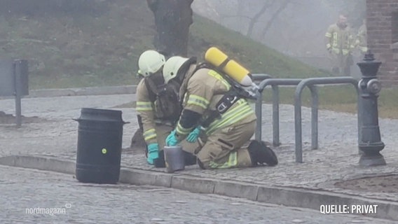 Zwei Einsatzkräfte der Feuerwehr mit Schutzmasken stellen einen Kanister sicher. © Screenshot 