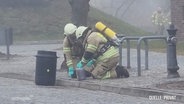 Zwei Einsatzkräfte der Feuerwehr mit Schutzmasken stellen einen Kanister sicher. © Screenshot 