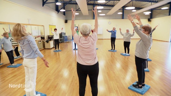 Seniorinnen stehen in einer Turnhalle im Kreis und strecken die Arme empor. © Screenshot 