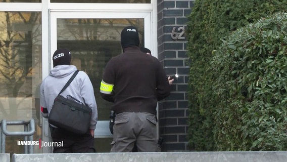 Vor einer Haustür eines größeren Wohnhauses stehen maskierte Spezialeinsatzkräfte der Polizei. © Screenshot 