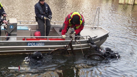 Feuerwehrtaucher heben ein verrostetes Fahrrad aus einem Alsterfleet auf ein kleines Boot. © Screenshot 