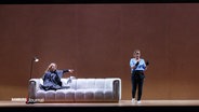 Ausschnitt einer Inszenierung der Hamburger Staatsoper: Eine Frau sitzt auf einem Sofa und zeigt auf eine andere Darstellerin. © Screenshot 