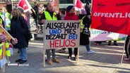 Streikende in Warnwesten protestieren auf dem Gänsemarkt. Zwei Männer halten ein Schild mit der Aufschrift "Ausgelaugt und mies bezahlt bei Helios". © Screenshot 