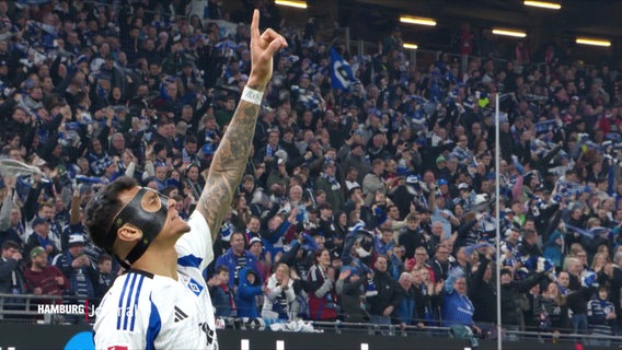 Davie Selke mit Gesichtsmaske und tätowiertem Arm zeigt jubelnd nach oben. Im Hintergrund feiernde HSV-Fans mit blau-weißen Schals im Stadion. © Screenshot 