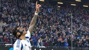 Davie Selke mit Gesichtsmaske und tätowiertem Arm zeigt jubelnd nach oben. Im Hintergrund feiernde HSV-Fans mit blau-weißen Schals im Stadion. © Screenshot 