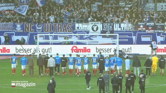 Die Holstein-Spieler in blauen Trikots stehen mit ihrem Team vor der Fankurve und blicken zu den jubelnden Fans mit Bannern und Fahnen im Stadion. © Screenshot 