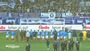 Die Holstein-Spieler in blauen Trikots stehen mit ihrem Team vor der Fankurve und blicken zu den jubelnden Fans mit Bannern und Fahnen im Stadion. © Screenshot 