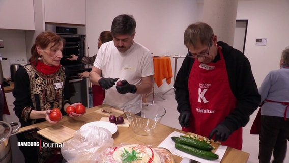 Menschen bereiten in einer Gemeinschaftsküche Gemüse zu. Ein Mann schneidet Gurken, ein anderer schält eine Zwiebel, während eine Frau Tomaten hält und spricht. © Screenshot 