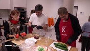 Menschen bereiten in einer Gemeinschaftsküche Gemüse zu. Ein Mann schneidet Gurken, ein anderer schält eine Zwiebel, während eine Frau Tomaten hält und spricht. © Screenshot 