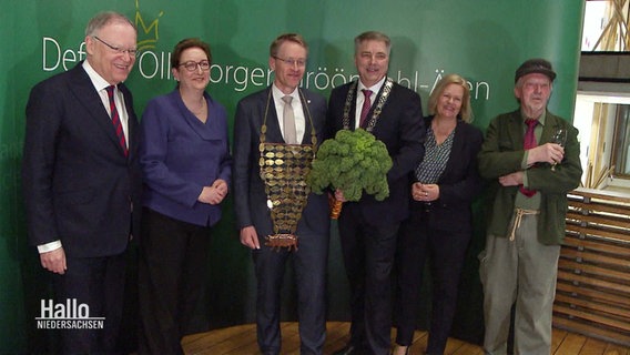 Daniel Günther (Mitte) steht mit Grünkohl in der Hand zwischen einer Reihe Frauen und Männer. © Screenshot 