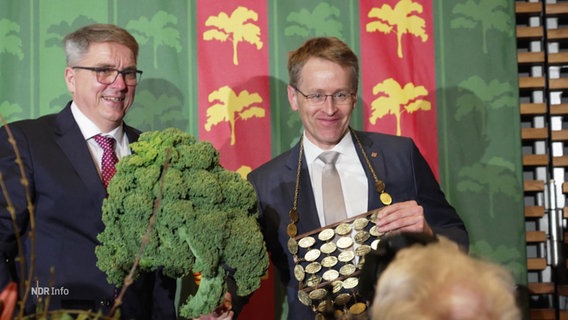 Daniel Günther lächelt in die Kamera und hält einen Grünkohl in der Hand. © Screenshot 