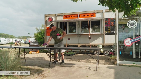 Neben einem Campingplatz steht ein Imbisswagen mit der Aufschrift "Larry's Grill Imbiss". Davor stehen Biertische, auf die der Betreiber gerade Blumentöpfe platziert. © Screenshot 