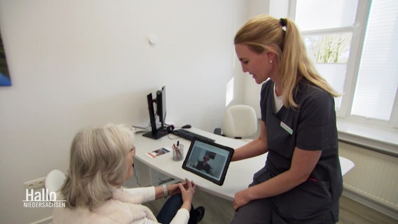 Eine sogenannte "Physical Assistant" sitzt mit einer Patientin in einem Behandlungsraum, ein Arzt ist auf einem Tablet zugeschaltet. © Screenshot 