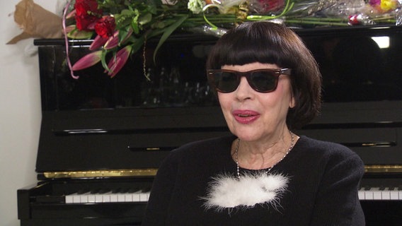 Mireille Mathieu mit dunkler Sonnenbrille sitzt vor einem Klavier. © Screenshot 