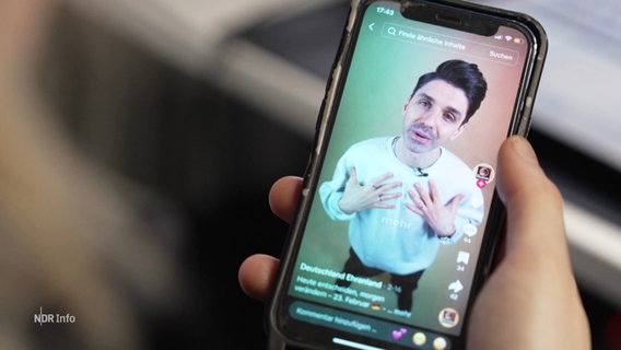 Eine Hand hält eine Handy, darauf läuft ein TikTok-Video von einem jungen Mann vor weißem Hintergund, der direkt in die Kamera spricht. © Screenshot 