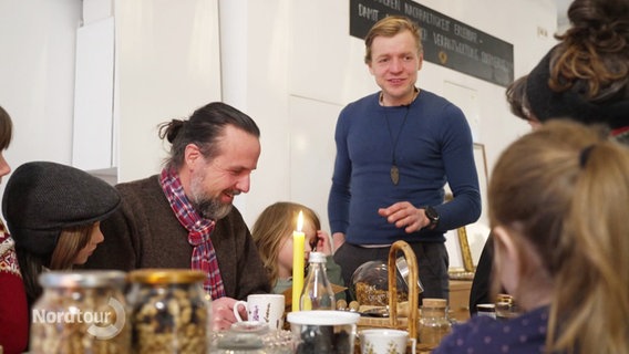 Cafébetreiber Robert von Leesen spricht zu mehrere Erwachsenen und Kindern, die an einem gedeckten Frühstückstisch sitzen. © Screenshot 