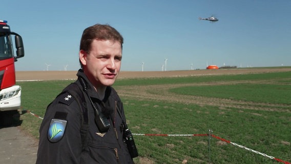 Moritz Horn von der Polizeihubschrauberstaffel Niedersachsen im spricht in die kamera. Im Hintergrund holt ein Hubschrauber Löschwasser aus einem Tank. © Screenshot 