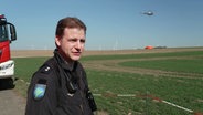 Moritz Horn von der Polizeihubschrauberstaffel Niedersachsen im spricht in die kamera. Im Hintergrund holt ein Hubschrauber Löschwasser aus einem Tank. © Screenshot 