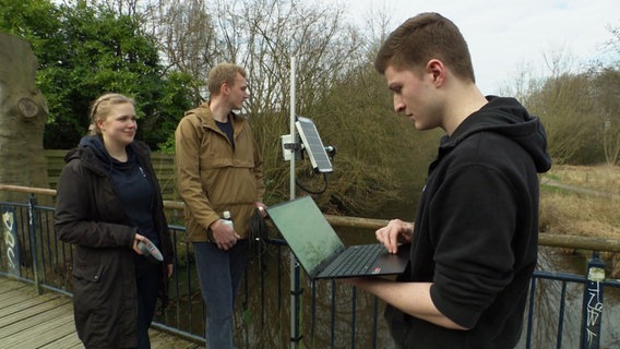 Eine Studentin und zwei Studenten stehen auf einer Brücke über einem Fluss. Ein Messgerät mit Solar-Panel ist am Geländer angebracht, einer der Studenten schaut auf einen Laptop. © Screenshot 