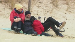 Eine Familie mit zwei kleinen Kindern sitzt zusammen am Strand. Sie alle tragen dicke Jacken, Mützen und Schals  