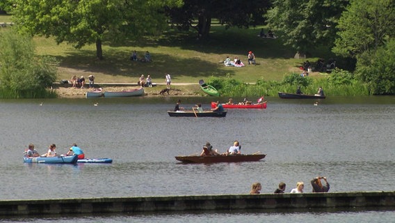 Verschiedene Boote auf einem kleinen See, im Hintergrund Spaziergänger.  