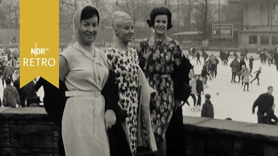 Drei Frauen posieren mit Kleidern vor einer Eisfläche mit Eisläufern  
