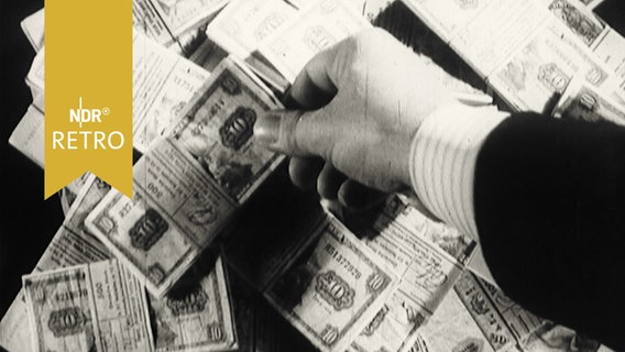 Eine Hand greift sich aus einem Haufen gebündelter DM-Scheine ein Bündel mit 10-Markscheinen heraus (1963)  