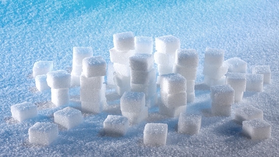 Loosely stacked sugar cubes on a surface with caster sugar.  © fotolia.com Photo: fotos4people
