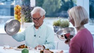 Doc Klasen und seine Patientin sitzen am gedeckten Tisch und lupfen die Deckel von den angerichteten Tellern. © NDR Foto: Moritz Schwarz/Oliver Zydek