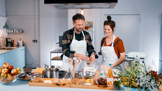 Ein Mann und Foodbloggerin Mia stehen mit Schürzen in der Küche und rühren einen Teig an. © NDR/Moritz Schwarz 