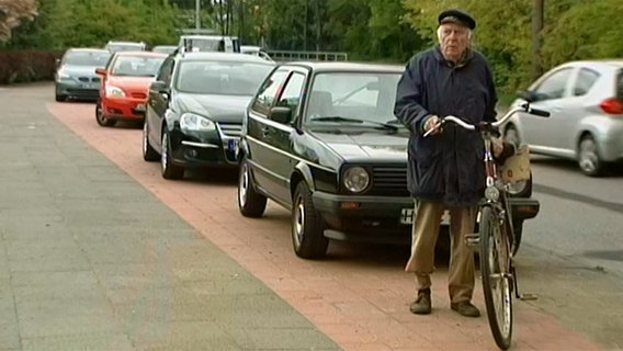 Mit Autos vollgeparkter Radweg, davor steht ein älterer Herr neben seinem Fahrad  