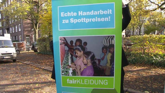 Ein Plakat wirbt mit der Aufschrift "Echte Handarbeit zu Spottpreisen" für Kleidung, die in Kinderarbeit hergestellt wurde.  
