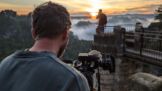 Ein Kameramann filmt eine Person im Hintergrund, die auf einer von Nebel umhüllten Aussichtsplattform im Gebirge steht. © NDR Foto: Maximilian Pilz