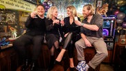 Guido Maria Kretschmer, Ina Müller, Bettina Tietjen und Sebastian Ströbel sitzen bei Inas Silvesternacht auf dem Tresen des "Schellfischpostens" und prosten einander zu. © NDR/Morris Mac Matzen Foto: Morris Mac Matzen