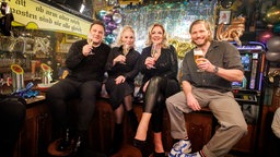 Guido Maria Kretschmer, Ina Müller, Bettina Tietjen und Sebastian Ströbel sitzen bei Inas Silvesternacht auf dem Tresen des "Schellfischpostens" und prosten in die Kamera. © NDR/Morris Mac Matzen Foto: Morris Mac Matzen