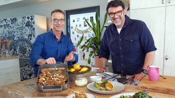 Fernsehkoch Tarik Rose und Ernährungs-Doc Matthias Riedl stehen in einer Küche und zeigen einen Teller mit Fischfrikadellen und eine Auflaufform mit Apple Crumble. © NDR/dmfilm/Florian Kruck 