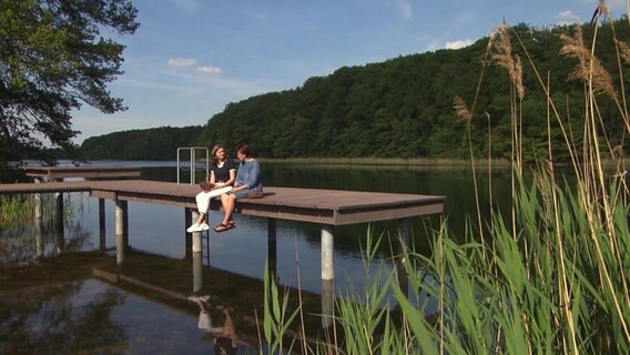 Heike Götz sitzt mit einer Frau auf einem Steg am See. © NDR 