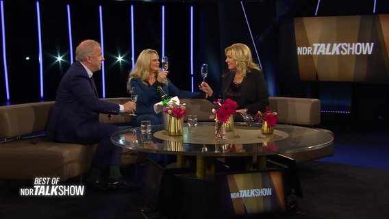 Die Moderatoren der NDR Talk Show, Barbara Schöneberger, Hubertus Meyer-Burckhardt und Bettina Tietjen sitzen im Studio und stoßen miteinander an. © NDR Screenshots 