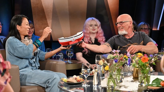 Björn Both, Frontmann der Band Santiano, reicht der Schauspielerin Minh-Khai Phan-Thi in der NDR Talk Show ein Spenden-Schiffchen der DGzRS hinüber und sie wirft eine Münze ein. © NDR Fernsehen/ Uwe Ernst Foto: Uwe Ernst