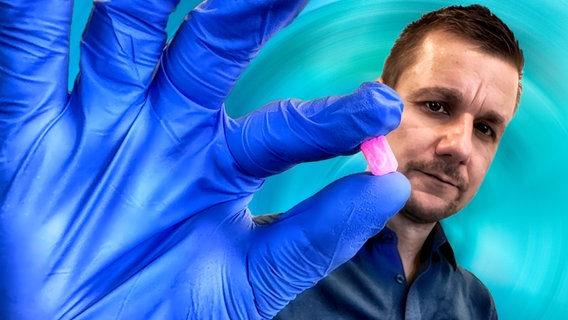 Moderator Tobi Schlegl hat blaue OP-Handschuhe an und hält mit der rechten Hand eine rechteckige, rosafarbene Ecstasy-Tablette in die Kamera. © NDR/Doclights | Look: Ulla Brauer 