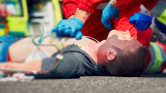 Aufnahme auf der Ebene eines Mannes mit nacktem Oberkörper, der gerade eine Herz-Druck-Massage von einem Rettungshelfer bekommt. Hinter einem Bereich mit Weichzeichner sind Schläuche zu erkennen. © SWR 